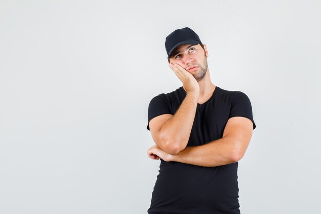 Delivery man in black t-shirt, cap leaning cheek on raised palm and looking pensive