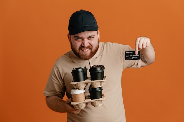 Free Photo delivery man employee in black cap and blank tshirt uniform holding coffee cups showing credit card looking at camera happy and positive smiling cheerfully standing over orange background