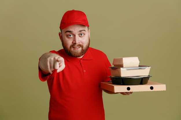 Free photo delivery man employee in red cap blank tshirt uniform holding food containers and pizza box pointing with index finger at camera smiling friendly standing over green background