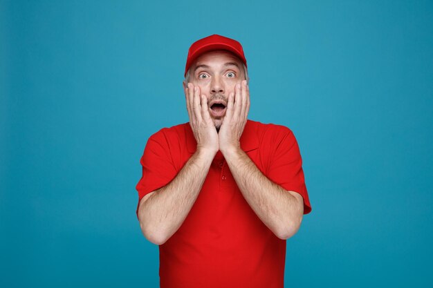 Delivery man employee in red cap blank tshirt uniform looking at camera amazed and surprised covering mouth with hands standing over blue background