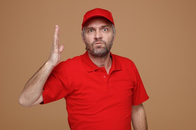 Free photo delivery man employee in red cap blank tshirt uniform looking at camera angry and frustrated raising arm in displeasure standing over brown background