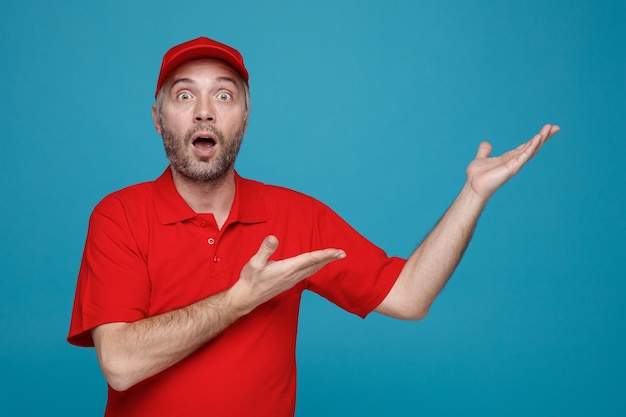 Delivery man employee in red cap blank tshirt uniform looking at cameraamazed and surprised presenting with arms of his hands something standing over blue background