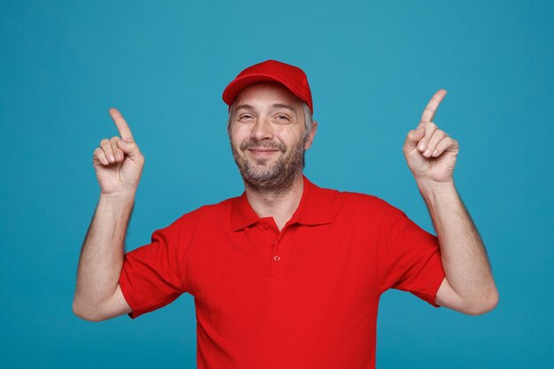 Delivery man employee in red cap blank tshirt uniform pointing with index fingers up smiling confident looking at camera standing over blue background