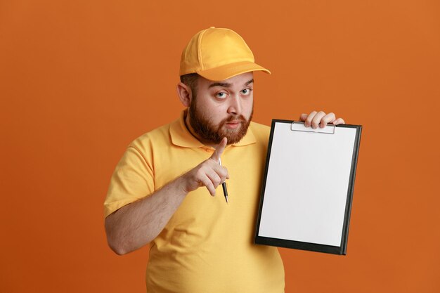 Delivery man employee in yellow cap blank tshirt uniform holding clipboard and pen pointing with index finger at camera being confident standing over orange background