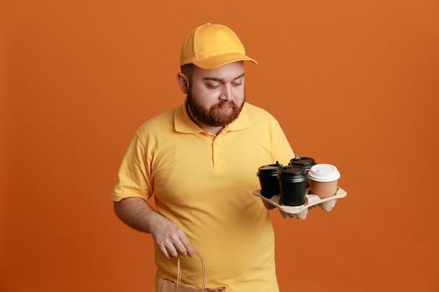 Free photo delivery man employee in yellow cap blank tshirt uniform holding coffee cups and paper bag looking at cups with serious face standing over orange background