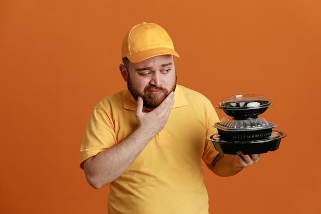 Delivery man employee in yellow cap blank tshirt uniform holding food containers looking at them with pensive expression thinking standing over orange background