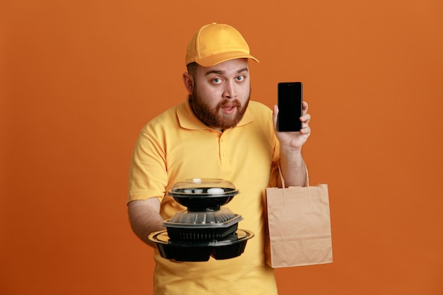 Delivery man employee in yellow cap blank tshirt uniform holding food containers with paper bag showing mobile phone looking at camera with serious face standing over orange background