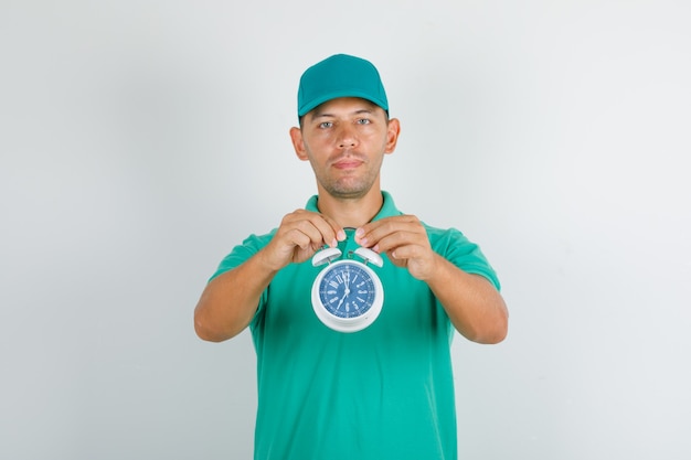 Free photo delivery man holding alarm clock in green t-shirt and cap and looking positive