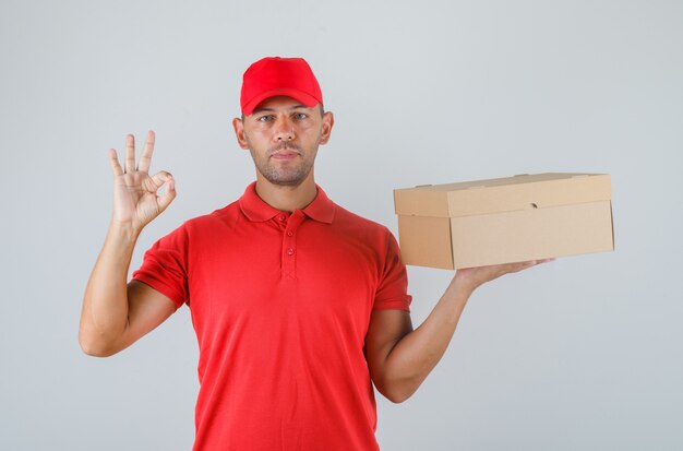 Delivery man holding cardboard box and showing ok sign in red uniform