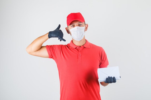 Delivery man holding envelopes and making call gesture in red uniform, medical mask, gloves , front view.