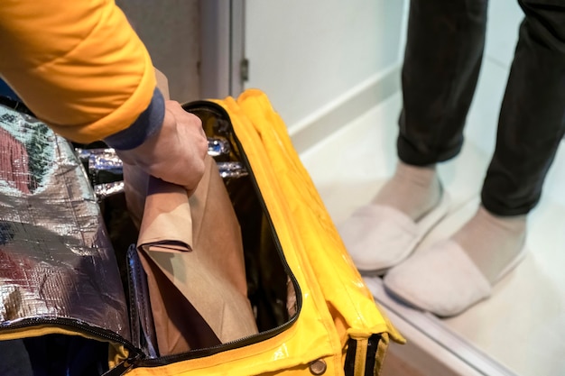 Delivery man opening yellow backpack and taking a bag with order, legs of other man