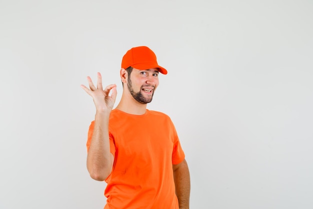 Delivery man in orange t-shirt, cap showing ok gesture and looking cheery , front view.