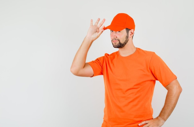 Delivery man posing with fingers on cap in orange t-shirt and looking confident. front view.