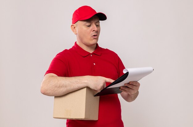 Delivery man in red uniform and cap holding cardboard box and clipboard looking at it with serious face standing over white background