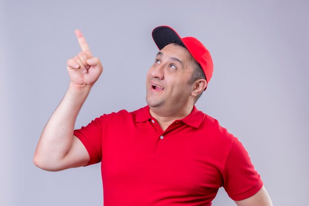 Delivery man in red uniform and cap smiling looking up and pointing with finger to something standing 