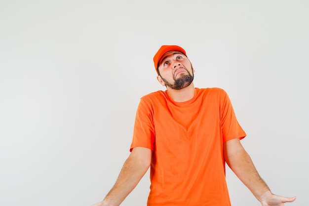 Delivery man showing helpless gesture by shrugging in orange t-shirt, cap and looking confused. front view.