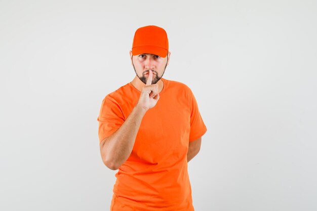 Delivery man showing silence gesture in orange t-shirt, cap and looking careful. front view.