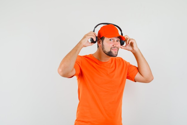 Free photo delivery man taking off headphones in orange t-shirt, cap and looking cheery , front view.