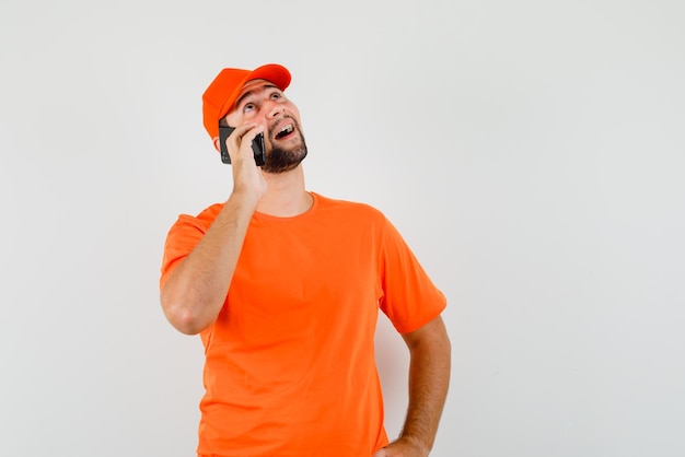 Delivery man talking on mobile phone in orange t-shirt, cap and looking hopeful , front view.