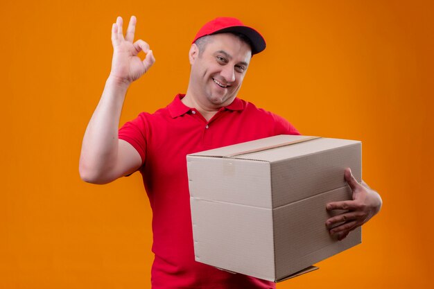 Delivery man wearing red uniform and cap holding box package looking positive and happy doing ok sign over orange wall