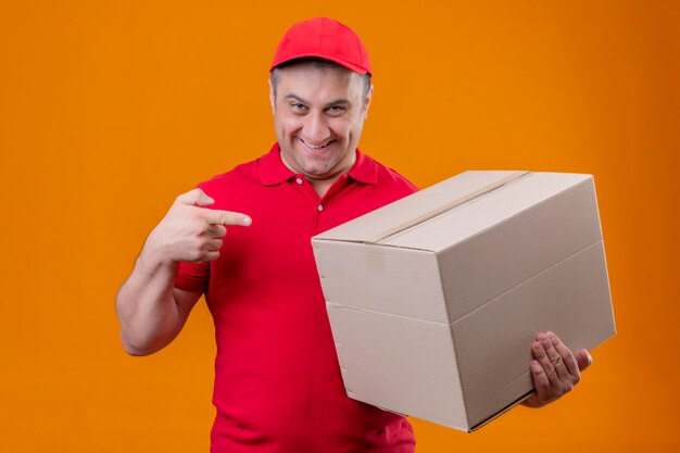 Delivery man wearing red uniform and cap holding large cardboard box pointing with index finger to it smiling confident over orange wall