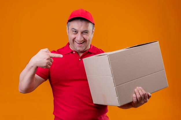 Delivery man wearing red uniform and cap holding large cardboard box pointing with index finger to it smiling confident over orange wall