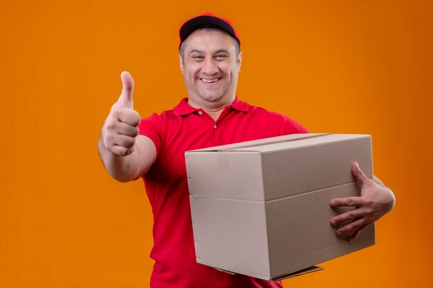 Delivery man wearing red uniform and cap holding paper package exited and happy raising fist after a victory standing over isolated blue space holding box package looking at came