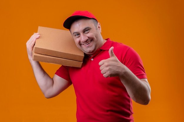 Free Photo delivery man wearing red uniform and cap holding pizza boxes positive and happy showing thumbs up over orange wall