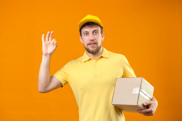 delivery man in yellow polo shirt and cap holding cardboard box smiling friendly doing ok sign standing on isolated orange