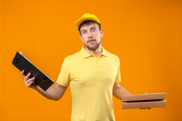 delivery man in yellow polo shirt and cap holding pizza boxes and clipboard looking at camera looking uncertain having doubts standing on isolated orange