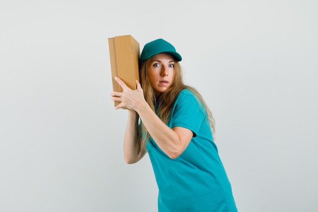 Delivery woman holding cardboard box near her ear in t-shirt, cap and looking curious. 