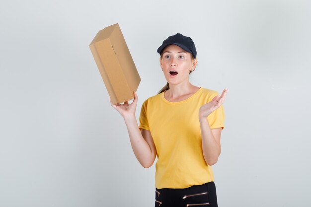 Delivery woman holding cardboard box in t-shirt, pants and cap and looking surprised