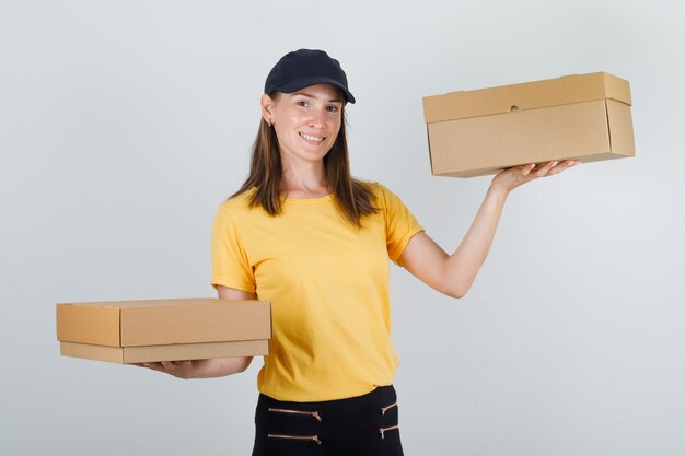 Delivery woman holding cardboard boxes in t-shirt, pants and cap and looking cheerful