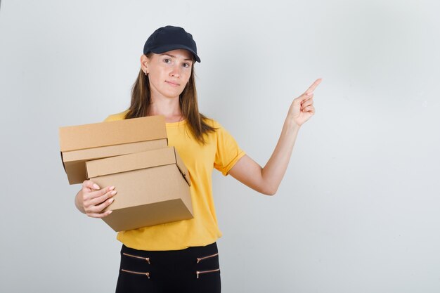 Delivery woman holding cardboard boxes with finger up in t-shirt, pants and cap