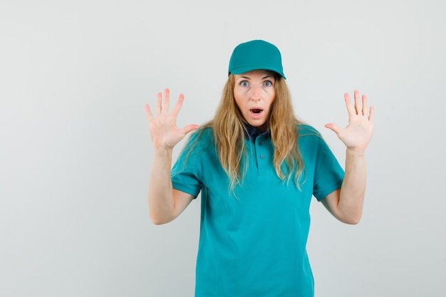 Delivery woman making surrender gesture in t-shirt, cap and looking scared 