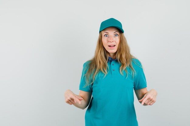 Delivery woman pointing down in t-shirt, cap and looking amazed 