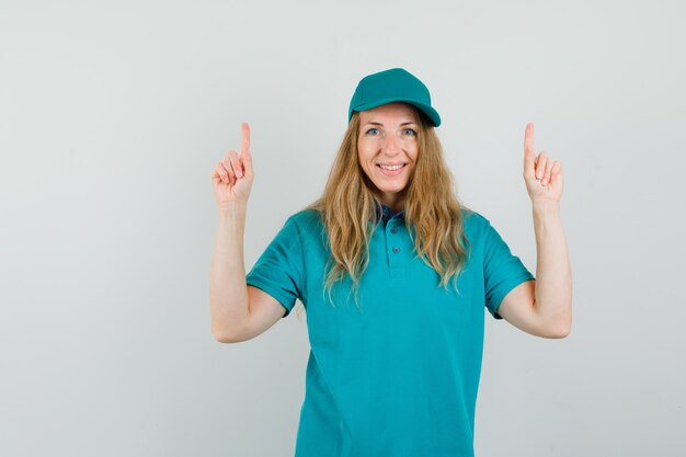 Delivery woman pointing up in t-shirt, cap and looking cheerful. 