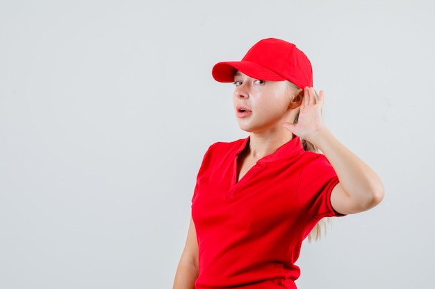 Free photo delivery woman in red t-shirt and cap keeping hand behind ear and looking curious