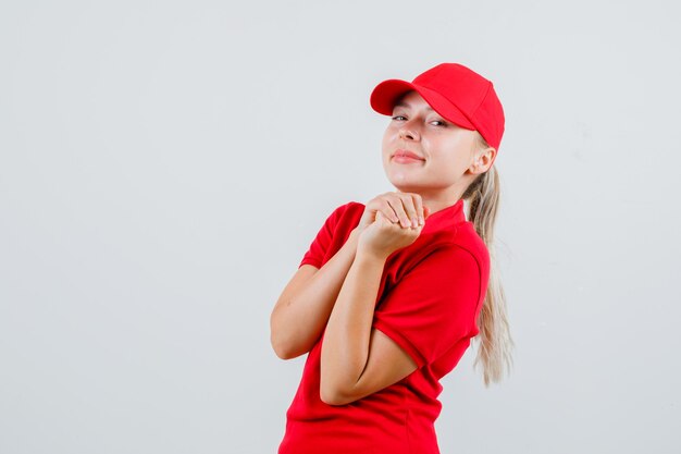 Delivery woman in red t-shirt and cap keeping hands clasped and looking merry