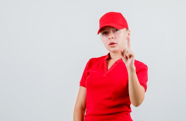 Free photo delivery woman in red t-shirt and cap pointing up and looking pensive