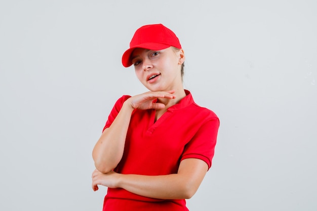 Free photo delivery woman in red t-shirt and cap propping chin on raised hand and looking cute