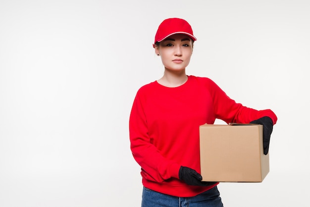 Delivery woman in red uniform isolated on white wall. Courier in medical gloves, cap, red t-shirt working as dealer holding cardboard box to deliver. Receiving package.