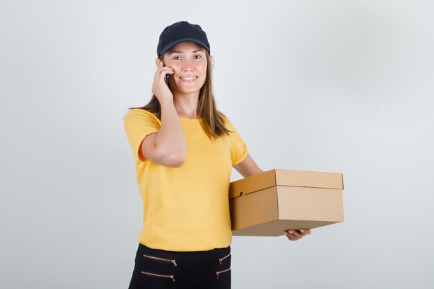 Delivery woman talking on smartphone with cardboard box in t-shirt, pants and cap