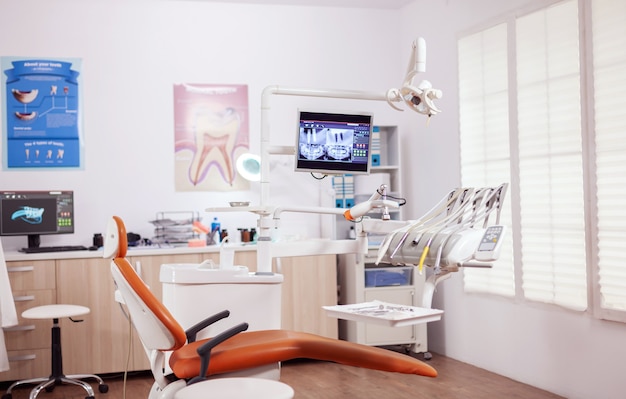 Dental chair and other accesorries used by dentist in empty cabinet. Stomatology cabinet with nobody in it and orange equipment for oral treatment.