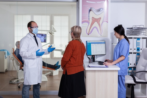 Dentist doctor with face mask explaining diagnosis to senior woman patient in stomatology waiting area hallway. Elderly man sitting on chair for teeth treatment.