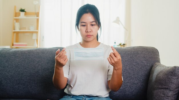 Depressed Asian business woman wearing protective mask sitting on sofa in living room at house when social distancing stay at home and self quarantine time, pandemic in china, coronavirus concept.