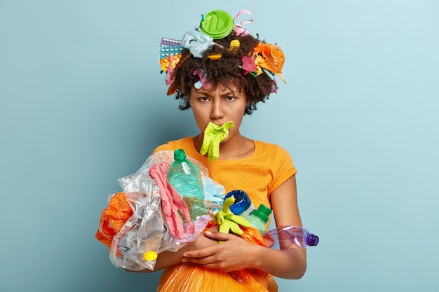 Free Photo depressed black woman with crisp hair, picks up garbage, has annoyed negative facial expression, cleans environment isolated over blue wall, sorts litter. people, recycling, volunteering concept