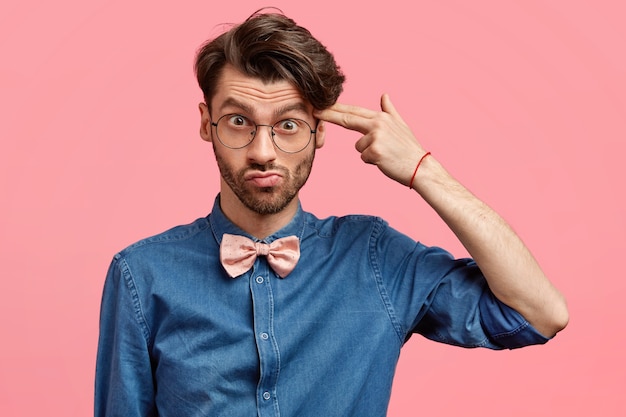 Depressed displeased unshaven male with trendy haircut, pretends killing himself, shoots with fore finger in temple, dressed in denim shirt and pink bowtie, feels tired of difficult life. Negativity