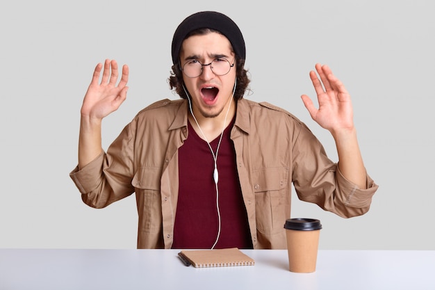 Depressed emotive guy raises hands and exclaims desperately, has annoyed facial expression, wears round optical spectacles, listens music, writes notes in notepad, isolated on white