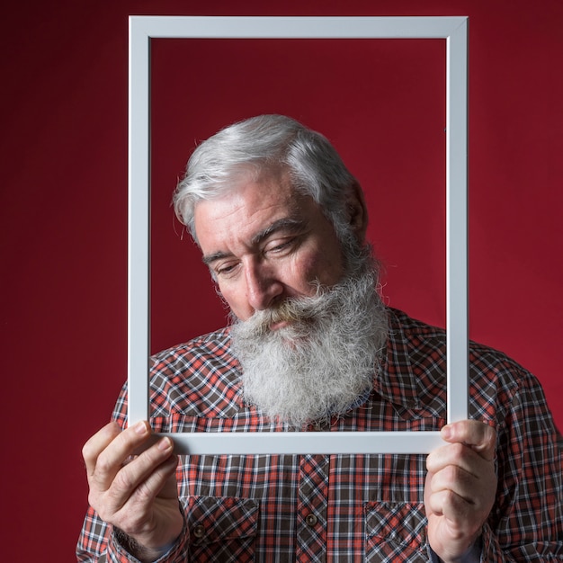 Free photo depressed senior man holding white border frame in front of his face against colored background